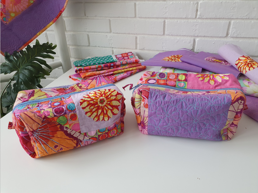 2 bags on a white table, one has a big embroidered flower in yellow and red, the other has a purple rectangle with quilted flower pattern. Some fabric scraps can be seen in the background.