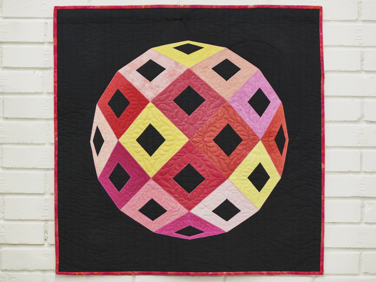 Two quilts are shown on the picture. One hanging on a ladder on the wall, the other is on a table to the right. Both quilts feature purple, pink, yellow, orange& green fabrics with embroidered flowers