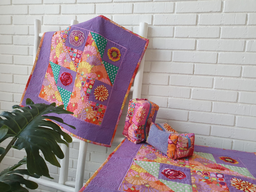 Two quilts are shown on the picture. One hanging on a ladder on the wall, the other is on a table to the right. Both quilts feature purple, pink, yellow, orange& green fabrics with embroidered flowers