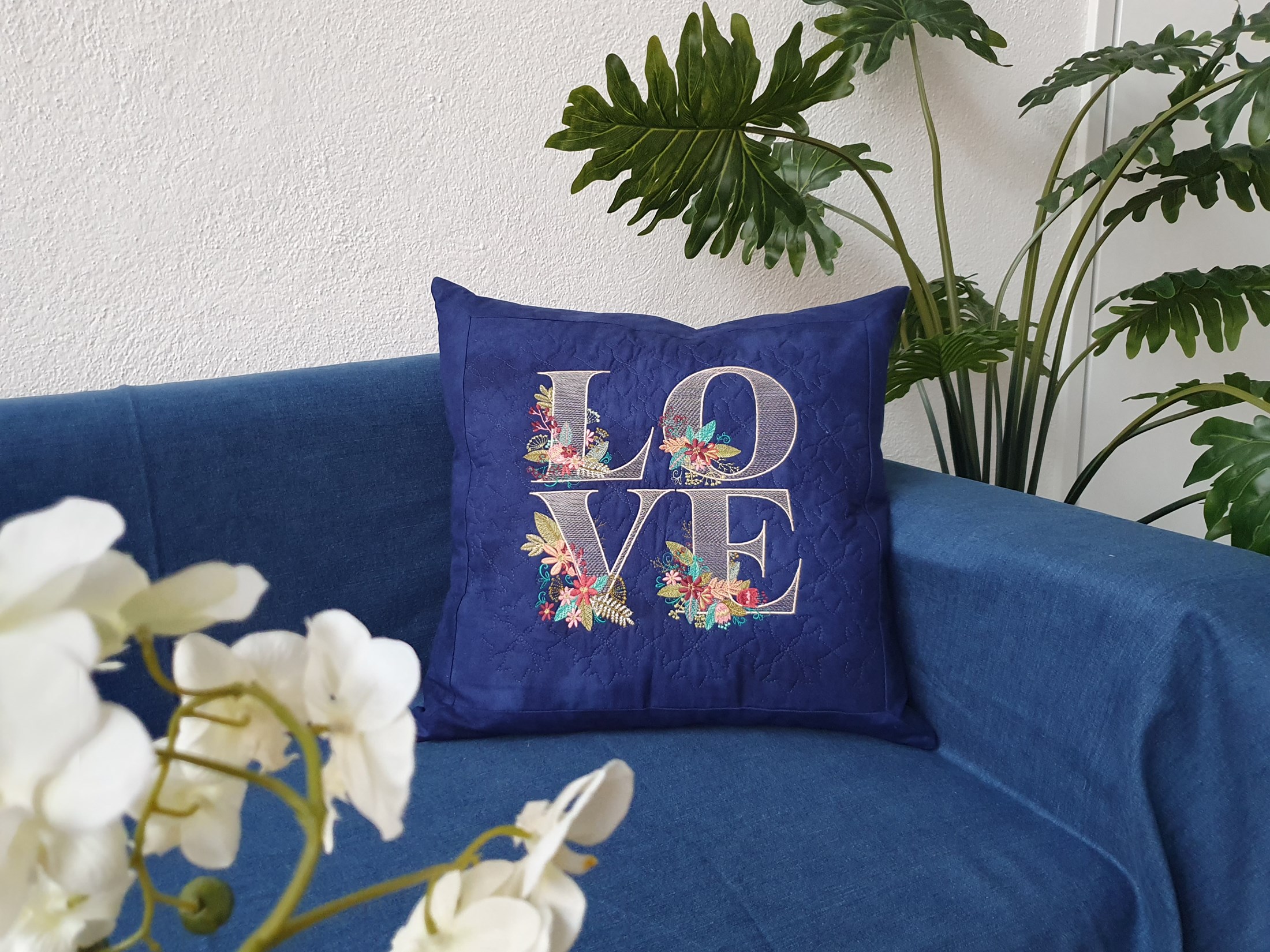A navy blue square pillow sits on a jeans-blue couch. There are white orchids on the left and a different plan on the right. The pillowcase is quilted and features an embroidery of the word LOVE.